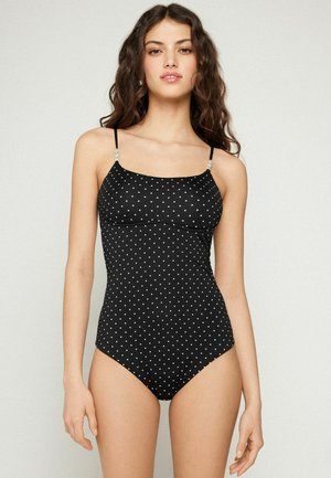 Mladá žena v černých jednodílných plavkách s bílými puntíky a úzkými nastavitelnými ramínky stojí před jednobarevným pozadím.