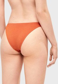 Femme portant un bas de bikini texturé orange, vue de dos, avec le bras détendu le long du corps sur un fond uni.