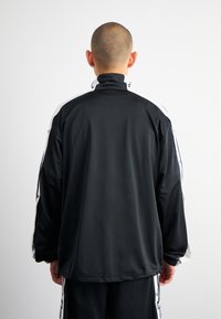 Svart träningsjacka med hög krage, långa ärmar och vita sidosträck. Tillverkad av ett mjukt tyg med en avslappnad passform.