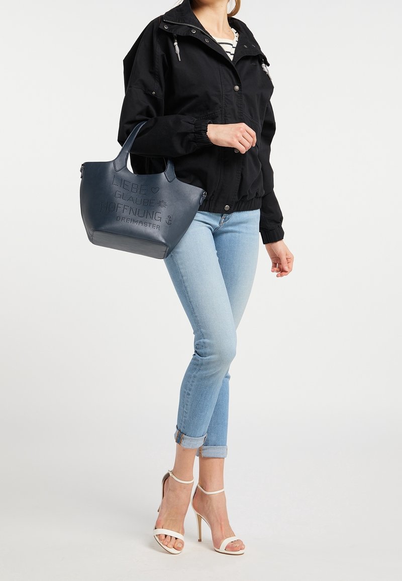 Femme portant une veste noire, un jean bleu clair retroussé, des sandales blanches à talons hauts, tenant un sac cabas bleu marine avec un texte en allemand.