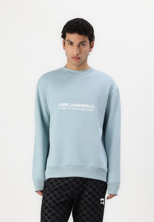 Sudadera azul claro con cuello redondo, mangas largas y logo impreso en blanco. Combinada con pantalones negros que presentan un patrón geométrico.