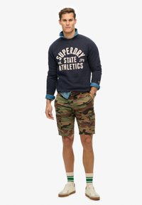 Felpa navy con testo bianco, camicia di jeans indossata sotto, pantaloni cargo mimetici, sneakers bianche e calzini a righe verdi. Abbigliamento casual.