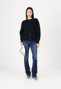 Schwarze Strickjacke mit Rundhalsausschnitt und großen Knöpfen, kombiniert mit blauen Jeans. Das Model hält eine silberne Clutch-Tasche und präsentiert einen lässigen Schnitt.