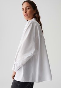 Vrouw met lang bruin haar, gekleed in een oversized witte blouse en zwarte broek, die over haar schouder kijkt tegen een effen achtergrond.