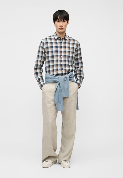 Άνδρας με πουκάμισο σε μπλε, καφέ και άσπρο καρό, μπεζ παντελόνι φαρδιάς γραμμής, λευκά αθλητικά παπούτσια, με ανοιχτό μπλε πουλόβερ δεμένο γύρω από τη μέση.