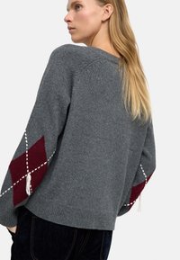 Szary dzianinowy sweter z szerokimi rękawami, ozdobiony bordowymi diamentowymi wzorami i białymi akcentami. Lekko luźny krój z prążkowanym dołem.