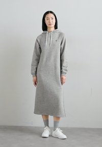 Grijze hoodie jurk gemaakt van zachte stof, met lange mouwen, een losse pasvorm en trekkoorddetails. Gecombineerd met witte sneakers.