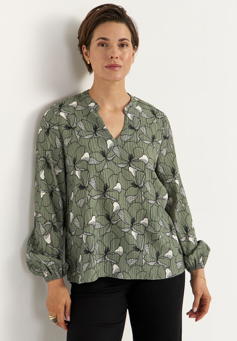 Luźna zielona bluzka z czarno-białym kwiatowym wzorem, dekoltem w serek oraz długimi rękawami zakończonymi elastycznymi mankietami. Lekka, fakturowana tkanina.