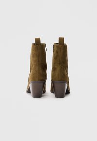 Botas de tobillo de ante verde oliva con un tacón apilado, costuras decorativas, cremalleras y lengüetas en la parte posterior.