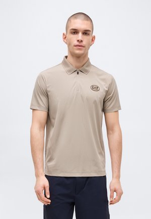 Ung mand med kort hår iført en beige kortærmet polo med frontlynlås og EA7-logo på brystet mod en ensfarvet baggrund.