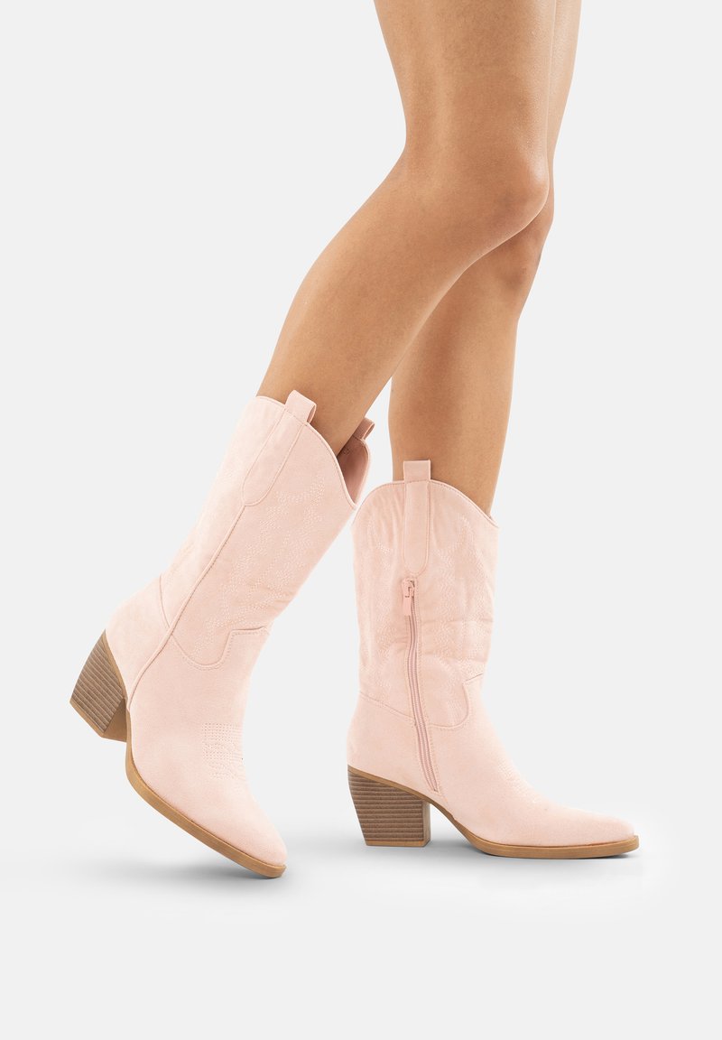 Różowe zamszowe kowbojskie botki z ostro zakończonym noskiem, średnim obcasem i bocznym suwakiem. Zawierają ozdobne przeszycia i lekko wygięty cholewkę.