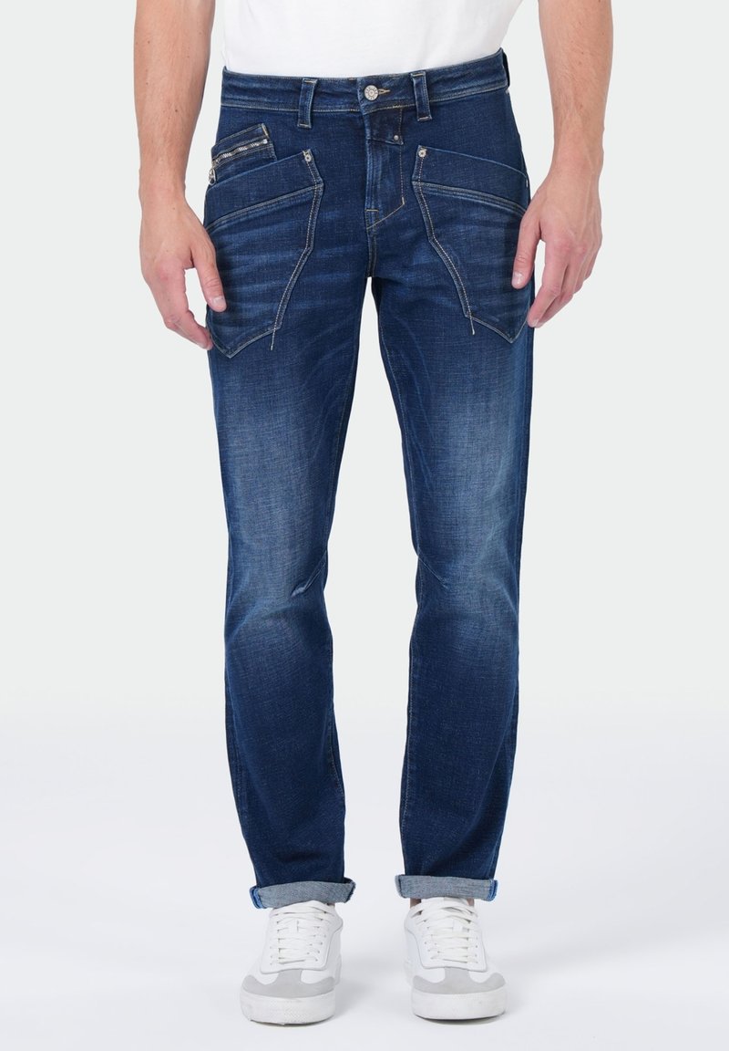 Donkerblauwe denim jeans met een slim fit, unieke ritssluiting en schuine zakken, en opgerolde manchetten. Gestyled met witte sneakers.