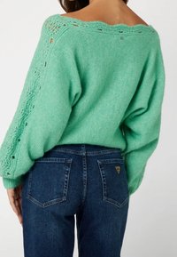 Pull en tricot vert avec un col large, des détails en dentelle et des manches texturées, associé à un jean en denim bleu avec des poches arrière et un logo.