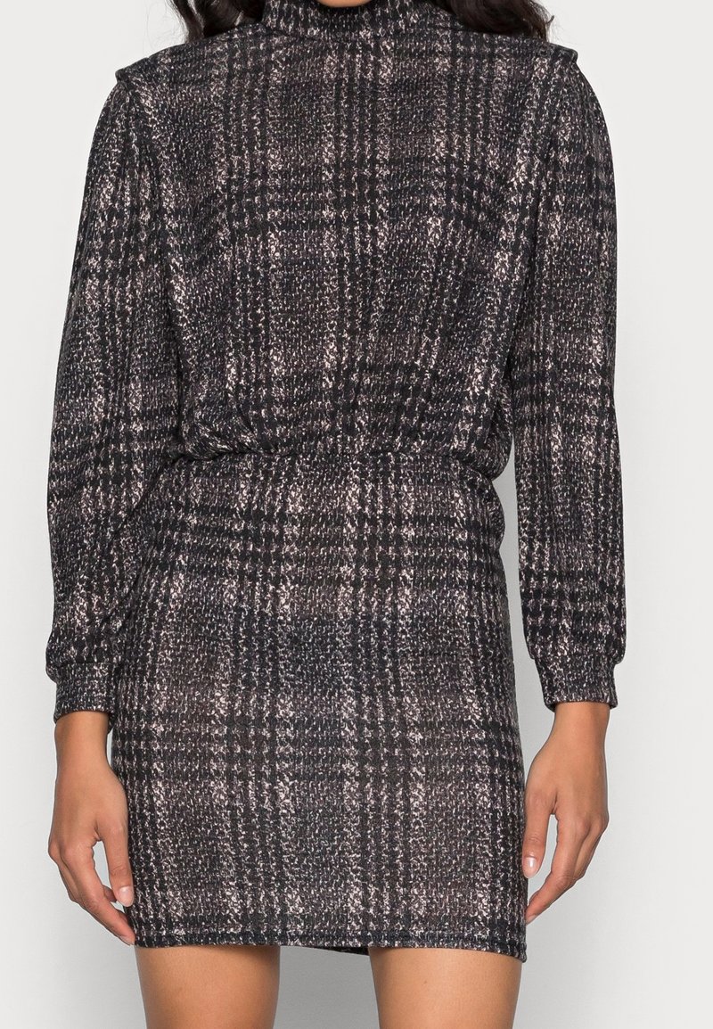 Femme portant une robe ajustée à manches longues et col montant avec un motif à carreaux noir et gris et une taille froncée.