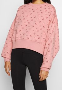 Rosa kortärmad sweatshirt med rund halsringning. Mönstrad med små röda cirklar och logotyper. Har långa, vida ärmar och en ribbad fåll.
