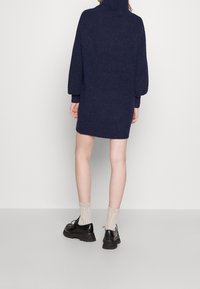 Mörkblå stickad klänning med hög krage, ballongärmar och ribbade manschetter, matchad med svarta skor och ljusbeige ribbade strumpor.