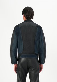 Vīrietis, tērpies melnā un tumši zilā džinsā, ar ādas biksēm, stāv ar muguru pret kameru uz vienkārša balta fona.