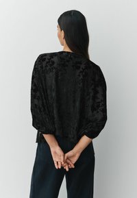 Black textured blouse with puffed sleeves, featuring a floral pattern. Worn with dark blue pants, hands clasped behind the back.