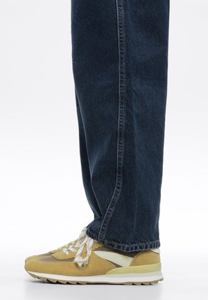 Zapatillas amarillas y beige con cordones blancos, usadas con vaqueros azul oscuro de corte recto sobre un fondo liso.