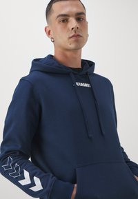 Hoodie bleu marine en coton doux, avec une poche frontale, capuche à cordon de serrage, présentant un logo blanc et des motifs en chevron sur les manches.