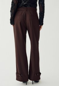 Smeđi krojeni hlače širokog kroja, s stražnjim džepovima i kopčama na manžetama. Glatka, čvrsta tkanina.