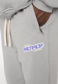 Pantalones de chándal grises con un tejido texturizado, que cuentan con un bolsillo con cremallera y un logo impreso en púrpura y blanco en el muslo izquierdo.
