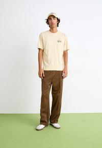 Beige cotton t-shirt with a green graphic on the chest, paired with brown wide-leg pants and white sneakers. Plain cap completes the outfit.