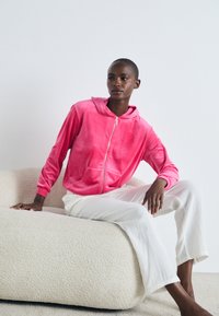 Suedine rosa com fecho éclair e bolso frontal, combinada com calças brancas com textura canelada, usada por uma pessoa sentada num sofá bege texturizado.