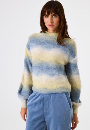 Woman wearing a pastel striped knit sweater with wave patterns and blue corduroy pants, standing against a white background.