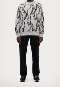 Sudadera gris con patrones de cadenas y texto en negro, con puños y dobladillo acanalados. Llevada con pantalones oscuros y zapatillas negras.