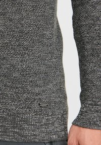 Jersey de punto en negro y gris con un patrón texturizado, que presenta puños y dobladillo de canalé, y una pequeña etiqueta de metal con logo en la parte inferior.