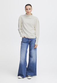 Haut crème à manches longues avec un col montant et des épaules bouffantes, associé à un jean en denim bleu à jambes larges et des baskets blanches.