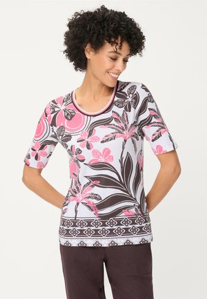 Lächelnde Frau mit lockigem Haar, die ein weißes Oberteil mit pinken und schwarzen Blumenmustern und eine dunkle Hose trägt, steht vor einem schlichten Hintergrund.