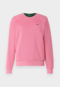 Różowa bluza z okrągłym dekoltem, prążkowanymi mankietami i paskiem dolnym. Na lewej piersi małe zielone logo Lacoste. Wykonana z teksturowanej tkaniny.