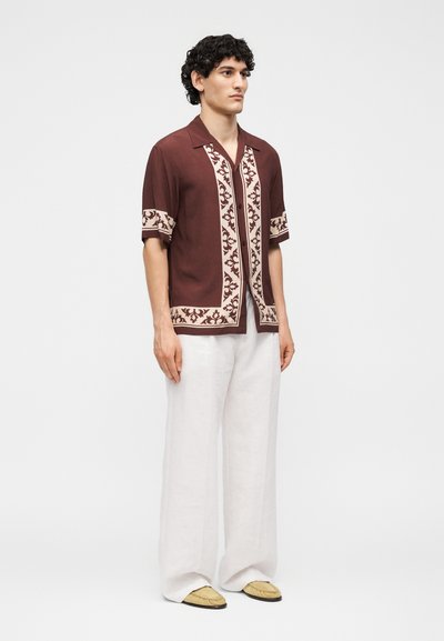 Άνδρας που στέκεται φορώντας καφέ κοντομάνικο πουκάμισο με μπεζ διακοσμητική ταινία, άνετο λευκό παντελόνι και μπεζ μοκασίνια σε απλό φόντο.