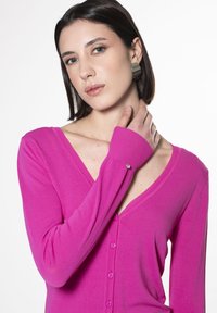 Cardigan a manica lunga con scollatura a V di un vivace colore rosa, con una texture a coste e chiusura a bottoni. Il polsino della manica è rifinito con un piccolo bottone decorativo.
