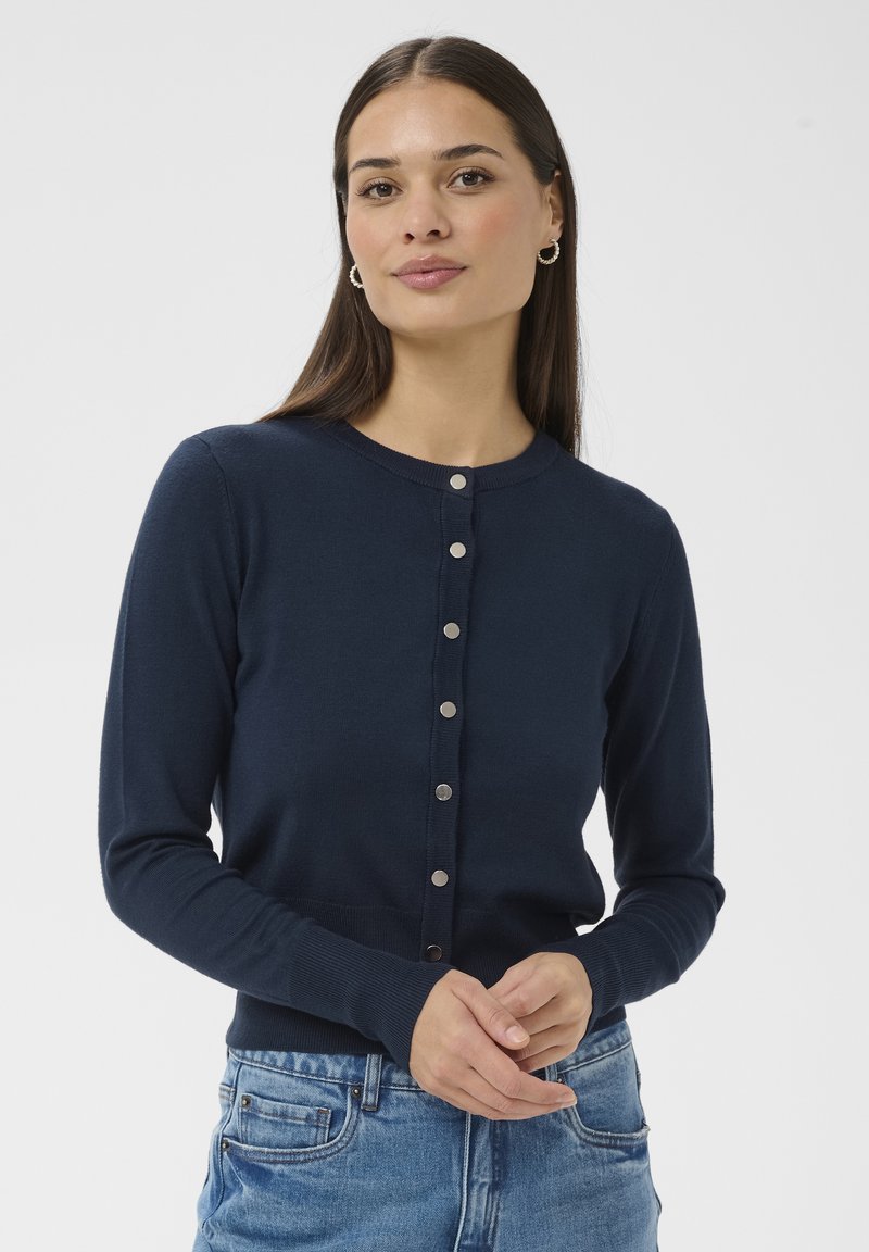 Femme aux longs cheveux foncés portant des boucles d'oreilles argentées en forme de créoles, un cardigan bleu marine boutonné et un jean bleu, debout devant un fond clair uni.