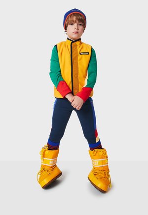 Farbige, mehrschichtige Jacke mit gelben, grünen und roten Ärmeln; marineblaue Hose; leuchtend gelbe Stiefel mit Schnürsenkeln; gestrickte Mütze.