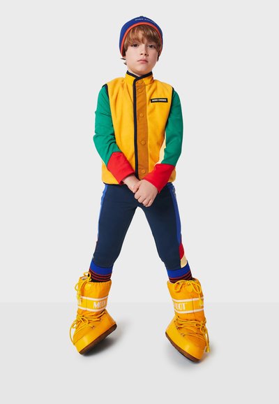 Färgglad, flerdimensionell jacka med gula, gröna och röda ärmar; marinblå byxor; ljusgula stövlar med snörning; stickad mössa.