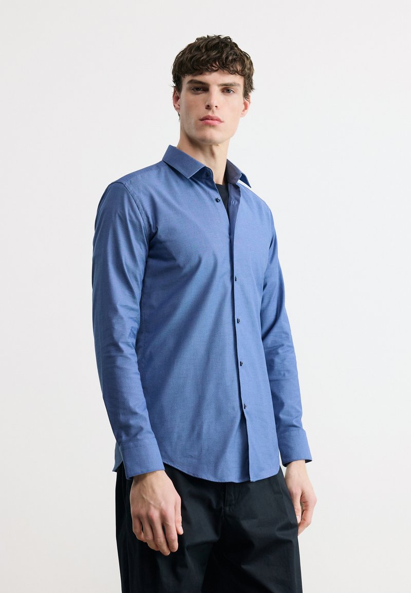 Άνδρας με κοντά, σγουρά μαλλιά φοράει στενή μπλε πουκάμισο με κουμπιά και μαύρο παντελόνι, στέκεται μπροστά σε λευκό φόντο.