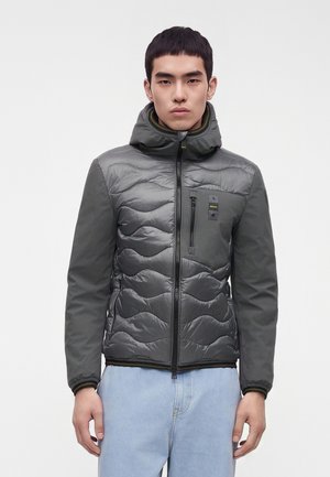 Ung mand iført en grå quiltet jakke med hætte og sort lynlås, parret med lyseblå jeans, står mod en ensfarvet baggrund.