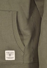 Olijfgroen sweatshirt met een grote voorzak, ribgebreide manchetten en zoom. Bevat een label met "TOP GUN" en een logo. Zachte textuur.