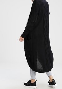 Cardigan nero lavorato a maglia con un design lungo e fluido, caratterizzato da un motivo a trecce sul retro e due tasche frontali.
