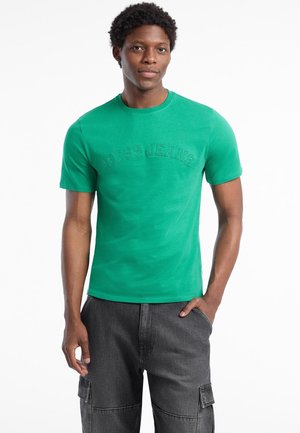 Green cotton t-shirt featuring embossed "GUESS JEANS" logo on the front, short sleeves, and a crew neck design. Paired with gray cargo shorts.
