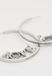 Boucles d'oreilles en argent en forme de créole ornées de lettrages décoratifs avec des accents floraux et une rangée de cristaux transparents le long du bord. Fini en métal lisse.