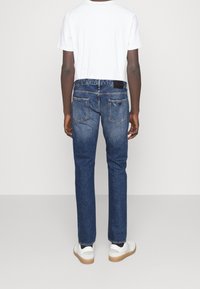 Modré džíny z denimu s rovnými nohavicemi, se dvěma zadními kapsami a černou nášivkou na pase, spárované s bílými teniskami.