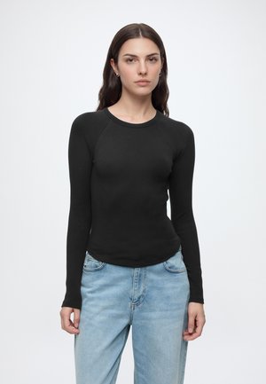 Giovane donna con capelli lunghi e scuri che indossa una maglia nera aderente a maniche lunghe e jeans azzurri, in piedi davanti a uno sfondo bianco uniforme.