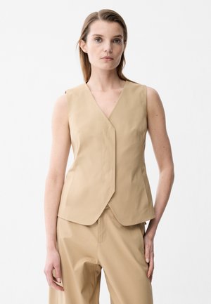 Kvinde med lys hud og lige brunt hår bærer en ærmeløs beige vest og matchende beige bukser mod en ensartet hvid baggrund.
