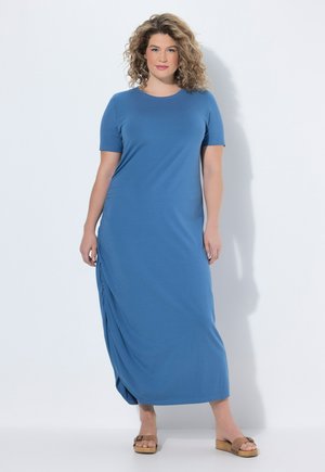 Femme aux cheveux bouclés portant une robe longue à manches courtes bleue et des sandales compensées beige, debout devant un fond clair.