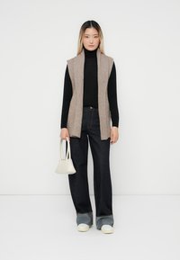 Gebreide beige vest met een ribbelstructuur, gedragen over een zwarte coltrui, gecombineerd met donkere flared jeans en grijze schoenen, met een kleine witte handtas.